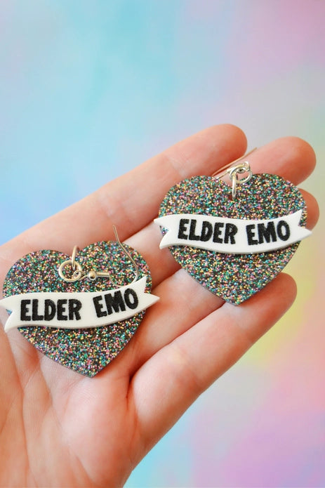 Earrings held in a White person's hand against a pastel pink and blue background