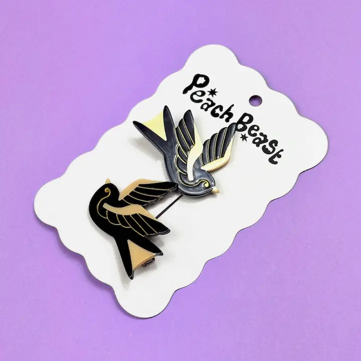 Set of two black and gold sparrow hair clips on a white card against a purple background.