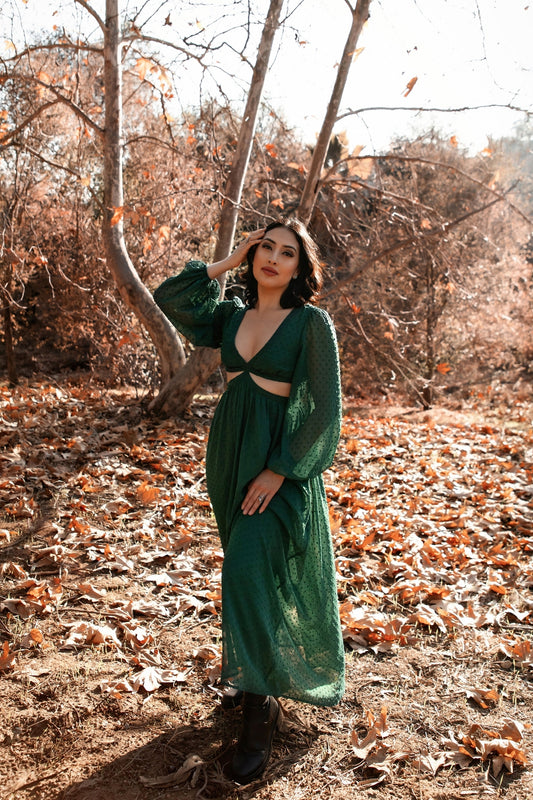 Female presenting model in the green Feralune dress standing in a forest with fallen leaves on the ground.