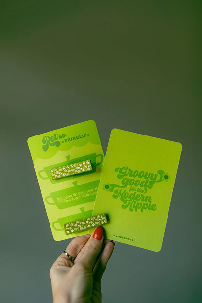 Model hand holding hair clips on a green display card against a green background.