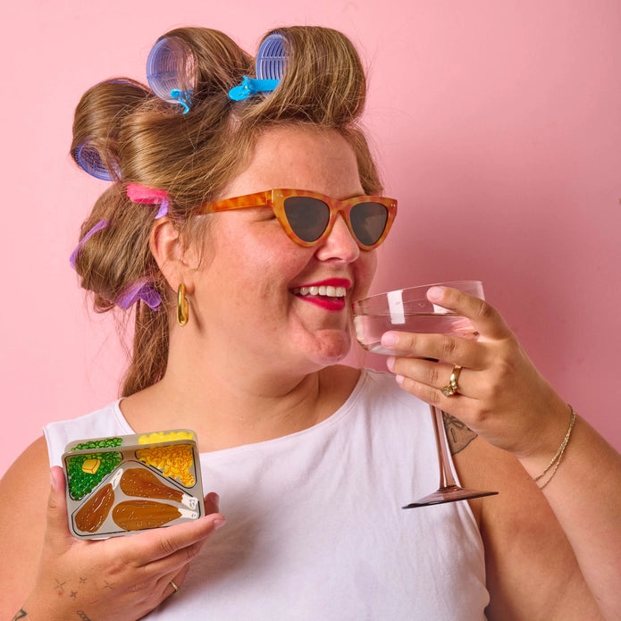 White model holding hair claw in one hand, and a beverage in another against a pink background.