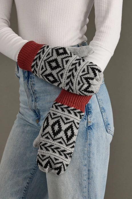 Person wearing patterned mittens with red cuffs and trim, paired with a white long-sleeve shirt and blue jeans.