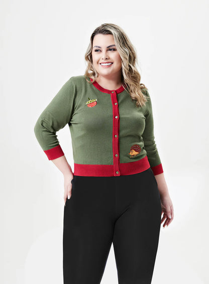 A plus-sized woman with wavy, shoulder-length blonde hair is smiling while wearing a light olive-green cardigan with bright red trim. The cardigan features playful embroidered patches, including a crowned watermelon slice on the left side of the chest and a red-tinted hedgehog on the lower right. The cardigan has three-quarter-length sleeves and is buttoned up the front. She pairs the cardigan with black pants against a white background.