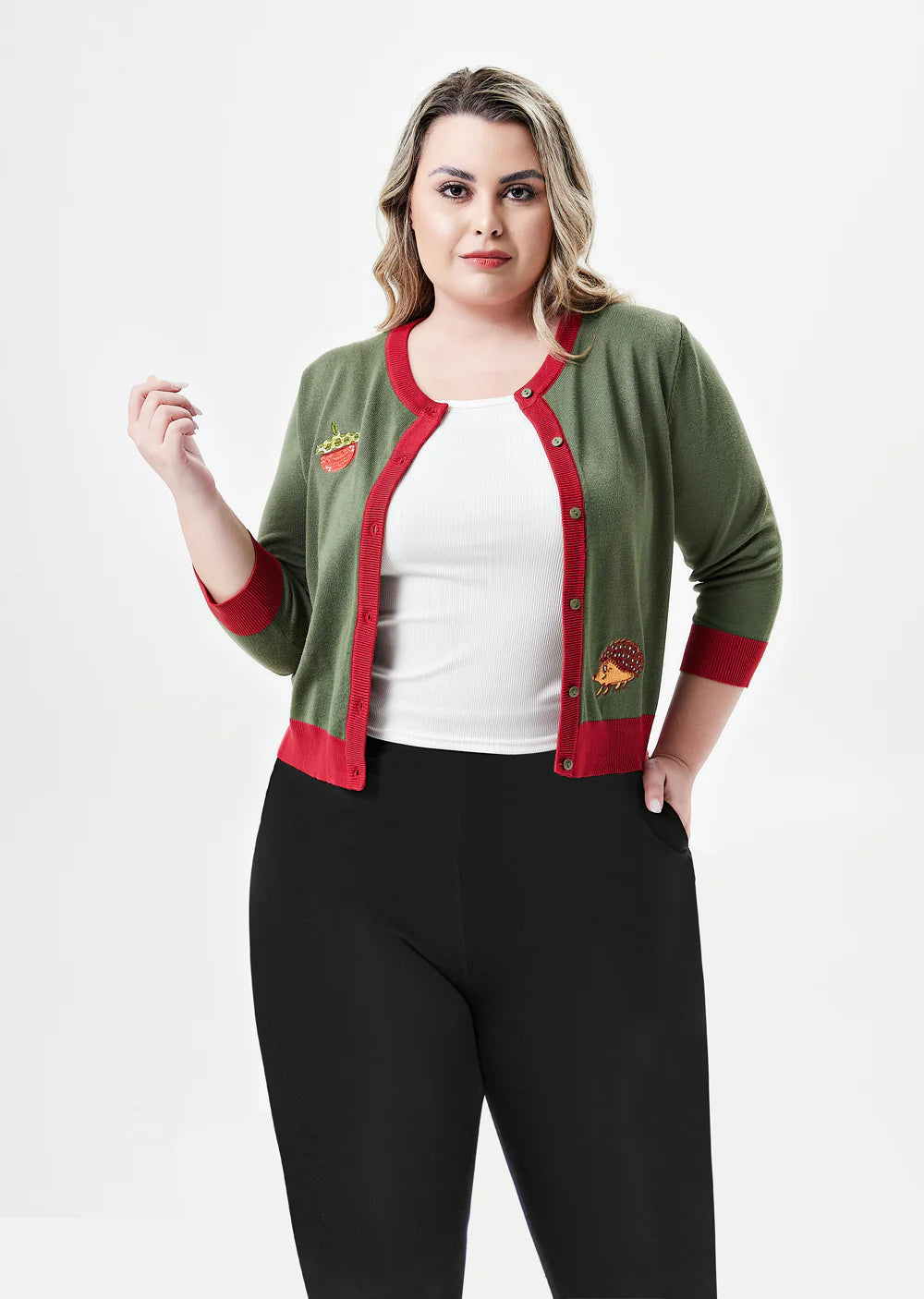 A plus-sized woman with wavy blonde hair is standing, facing forward with a neutral expression. She is wearing an olive-green cardigan with bright red trim and playful embroidered patches, including a crowned watermelon slice on the chest and a red-tinted hedgehog on the lower side. The cardigan is worn open over a white ribbed tank top and paired with black pants. The background is white, and her hands are casually resting in her pockets, with one hand lifted slightly.