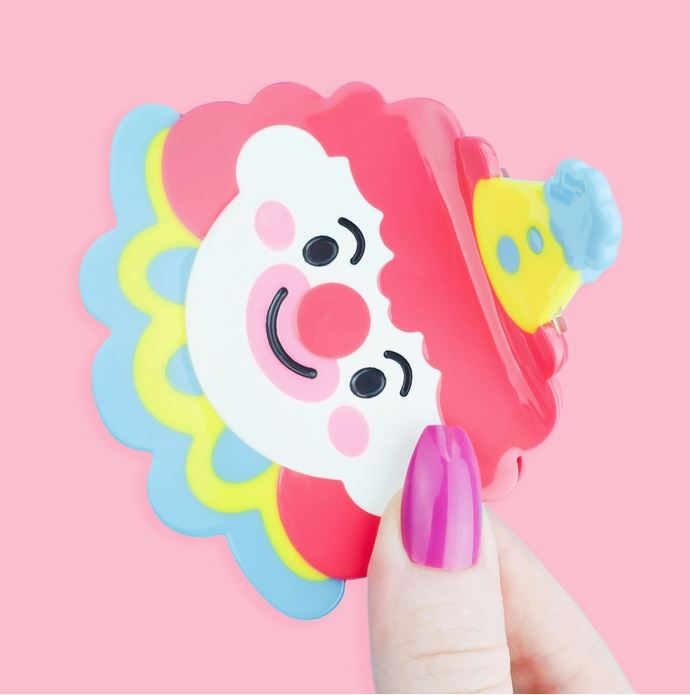 Clown with red hair with yellow and blue accessories as a hair claw against a pink background.