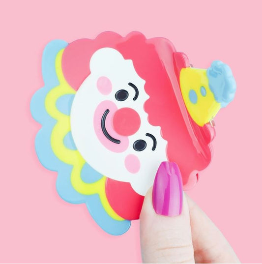 Clown with red hair with yellow and blue accessories as a hair claw against a pink background.