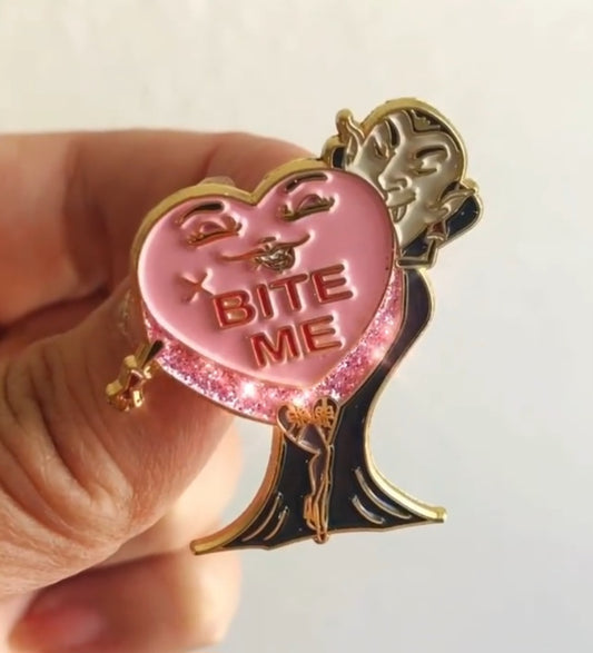 Pink heart with words: BITE ME in red. Heart has legs and arms and a face, and a vampire sinking their teeth into the heart. Pin is held by a hand against a white background