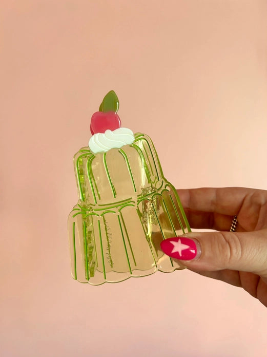 Transparent green jello cake hair claw with a cherry on top held by a hand against a pink background.