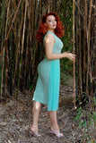 Model with bright red hair at a side angle towards the right with one leg back showcasing the side of the dress against a bamboo forest background