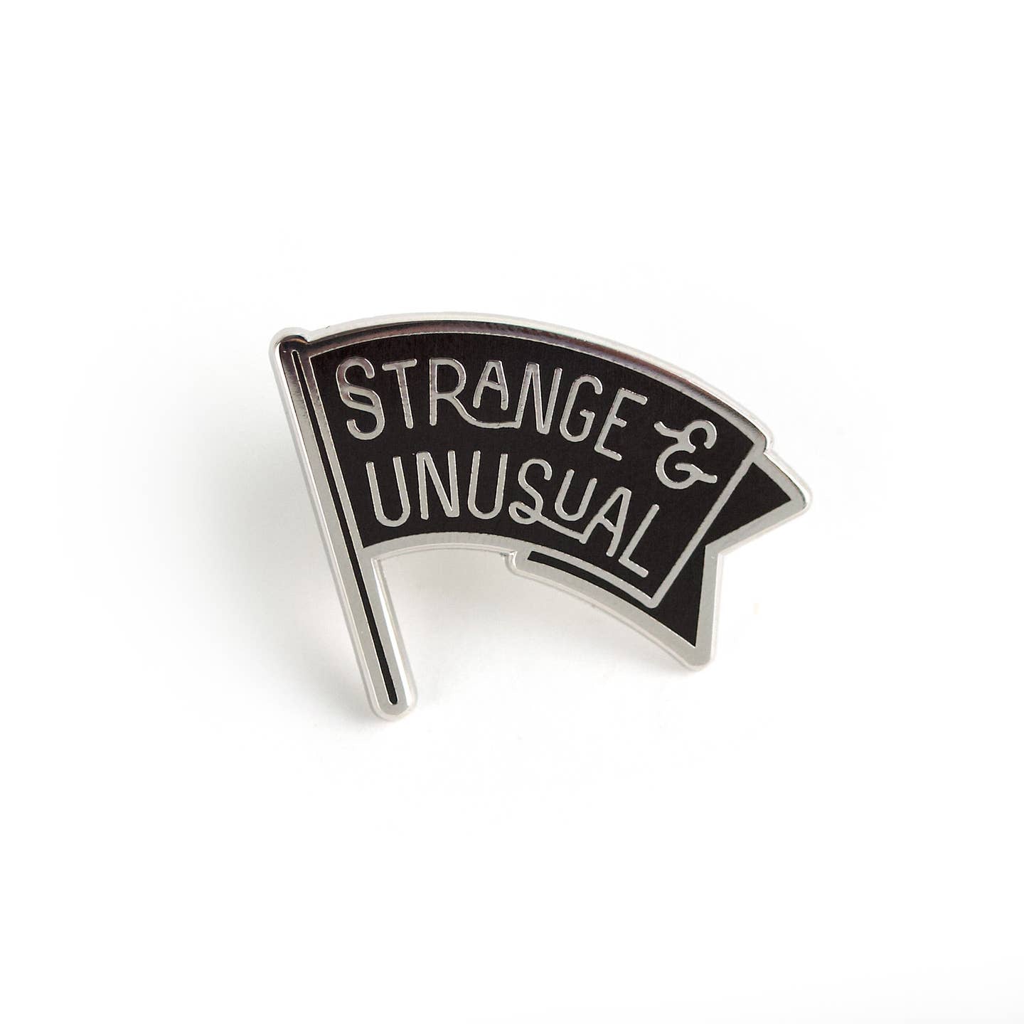 Pin is a black banner with a curve to it. Banner has silver letters that say "Strange & Unusual" instrad of using the word "and" it uses an ampersand. Banner has a thin silver outline around it. Pin is shown against a white background.