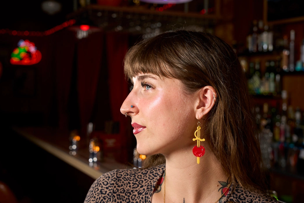 Woman wearing yellow sword earring with a cherry stabbed through it in a bar.