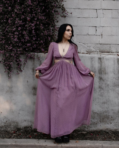 Female presenting model in the purple Lunara dress standing against a textured wall with plants.