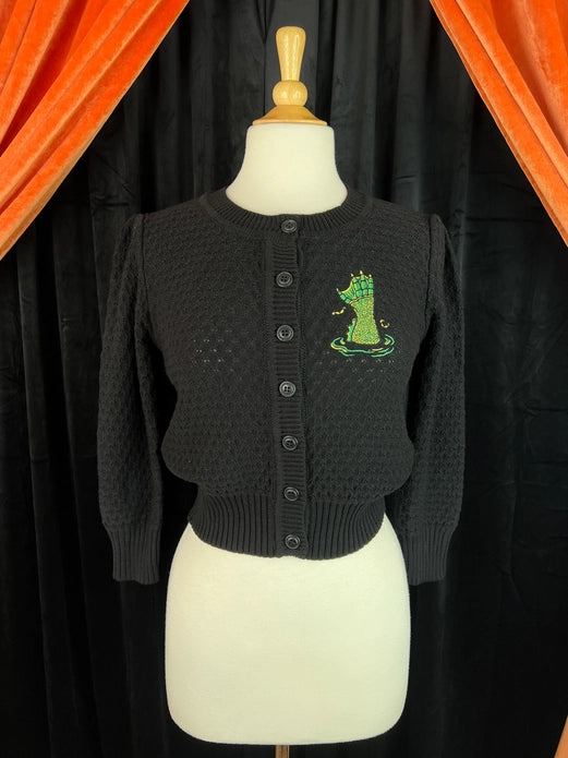 Black cardigan featuring an embroidered green Creature from the Black Lagoon inspired hand reaching upwards like out of water on the right side of the cardigan (left side when worn). Cardigan is on a mannequin with a black velvet background with orange drapery