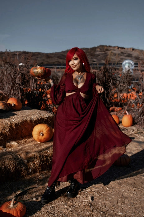 Woman in a burgundy dress standing in a pumpkin patch with hay bales.