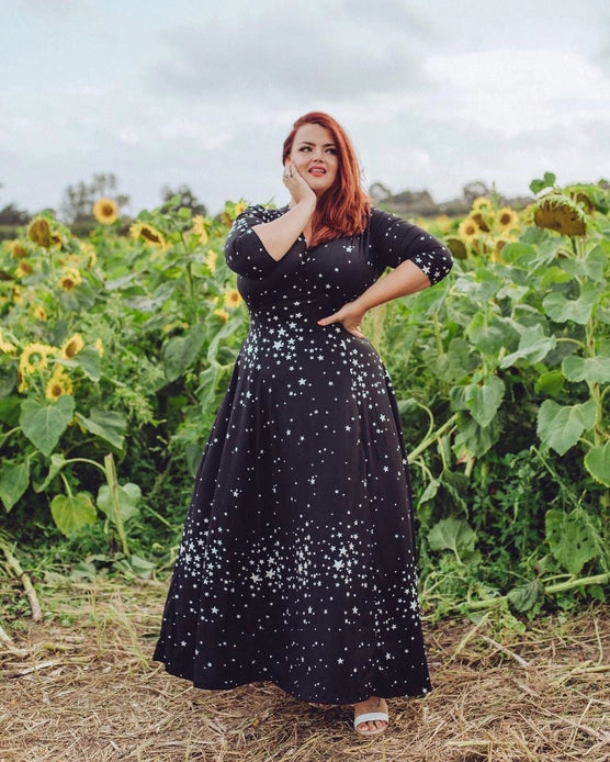 Model wearing dress showcasing the front of the dress with one hand on hip and other hand on face against a sunflower field