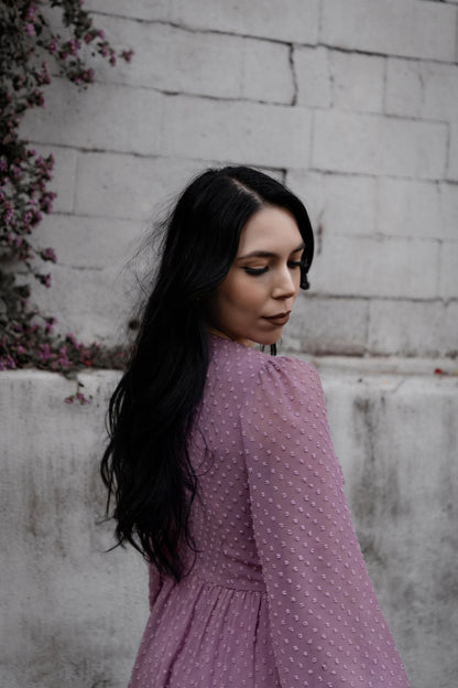 Female presenting model in the purple Lunara dress standing against a textured wall with plants.