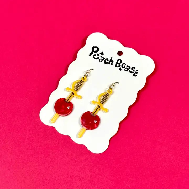 Yellow swords earrings with cherries stabbed through them on a white card against a pink background.