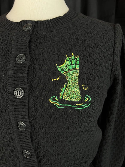 Close up of the embroidery that is on the left side of the cardigan. Embroidery features different shades of green mimicking the hand of The Creature from the Black Lagoon movie monster