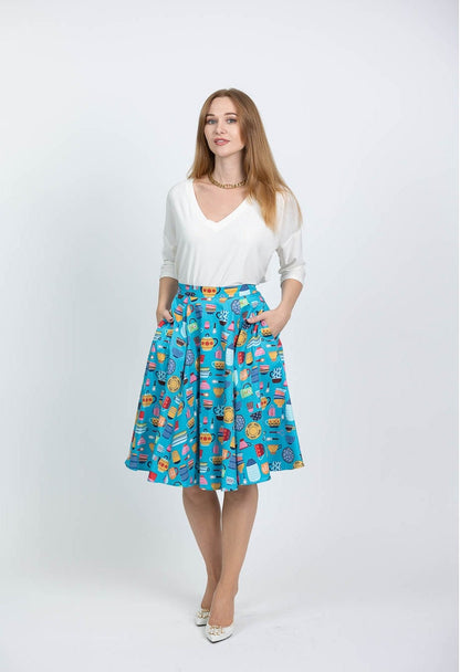 This photo shows a white female presenting model wearing a white blouse with the Kitchenware skirt. Skirt has a array of vintage kitchen items such as mixing bowls, tea kettles, mugs, etc on a light blue background.  Model is shown with hands in the pockets of the skirt