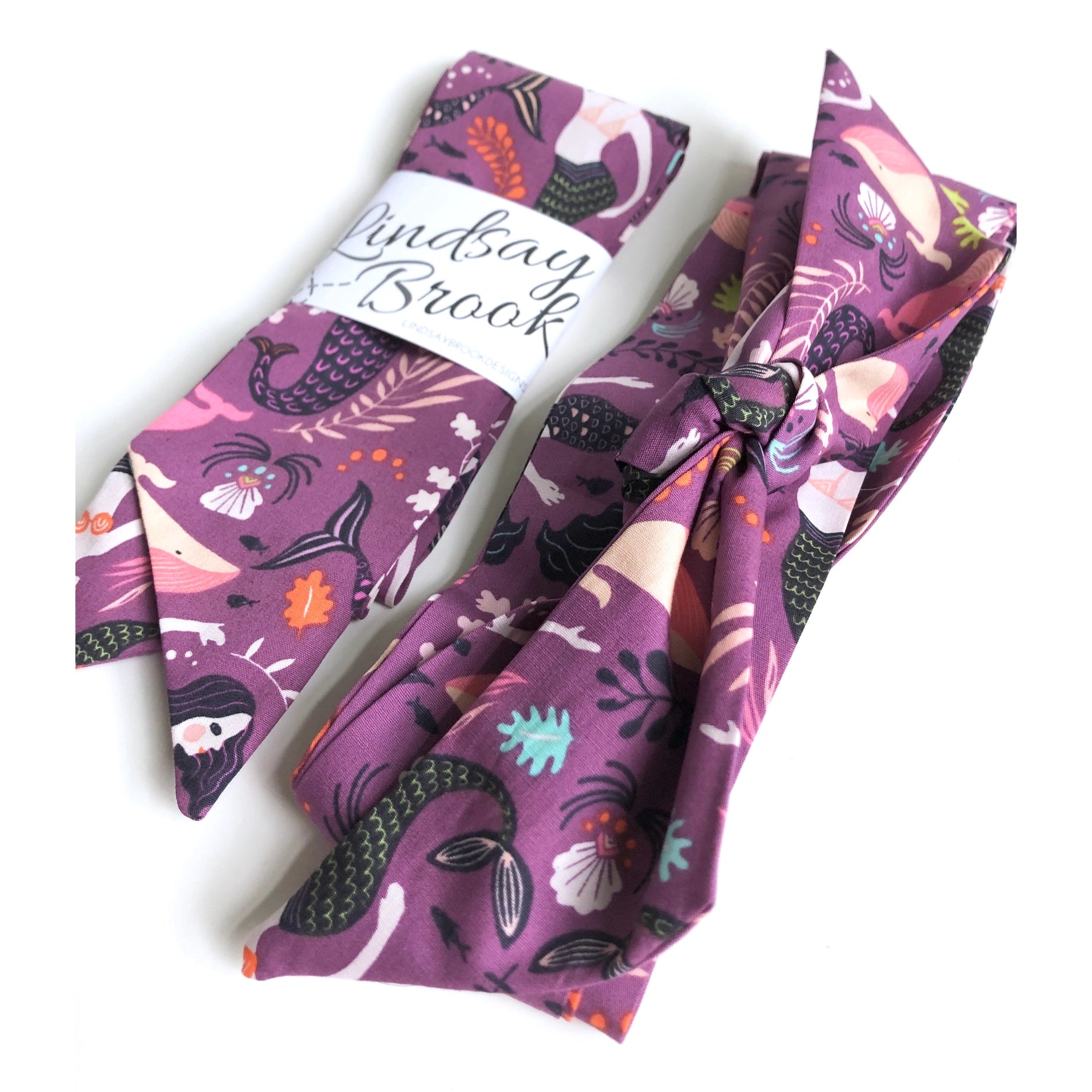 This photo showcases 2 hair scarves next to each other. One in the packaging, and one with a bow. This photo is at an angle. This scarf has different mermaids with dark colored tails, pink whales, and various sea plants on a dark purple background 