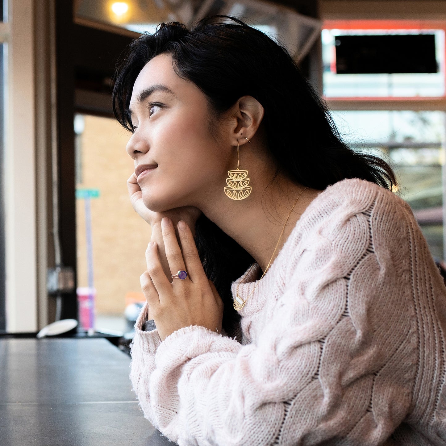 This photo shows a female presenting person wearing the earrings while staring out a window. Person is wearing a light pink sweater, and has dark hair