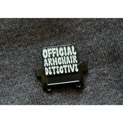 Photo shows an upclose look at the pin. Armchair is black with white lettering that says Official Armchair Detective. Pin is on a grey sweater