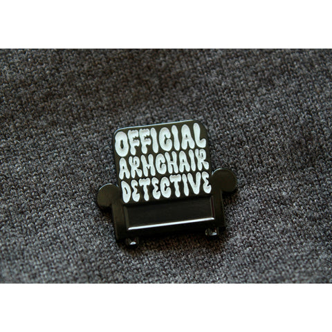 Photo shows an upclose look at the pin. Armchair is black with white lettering that says Official Armchair Detective. Pin is on a grey sweater