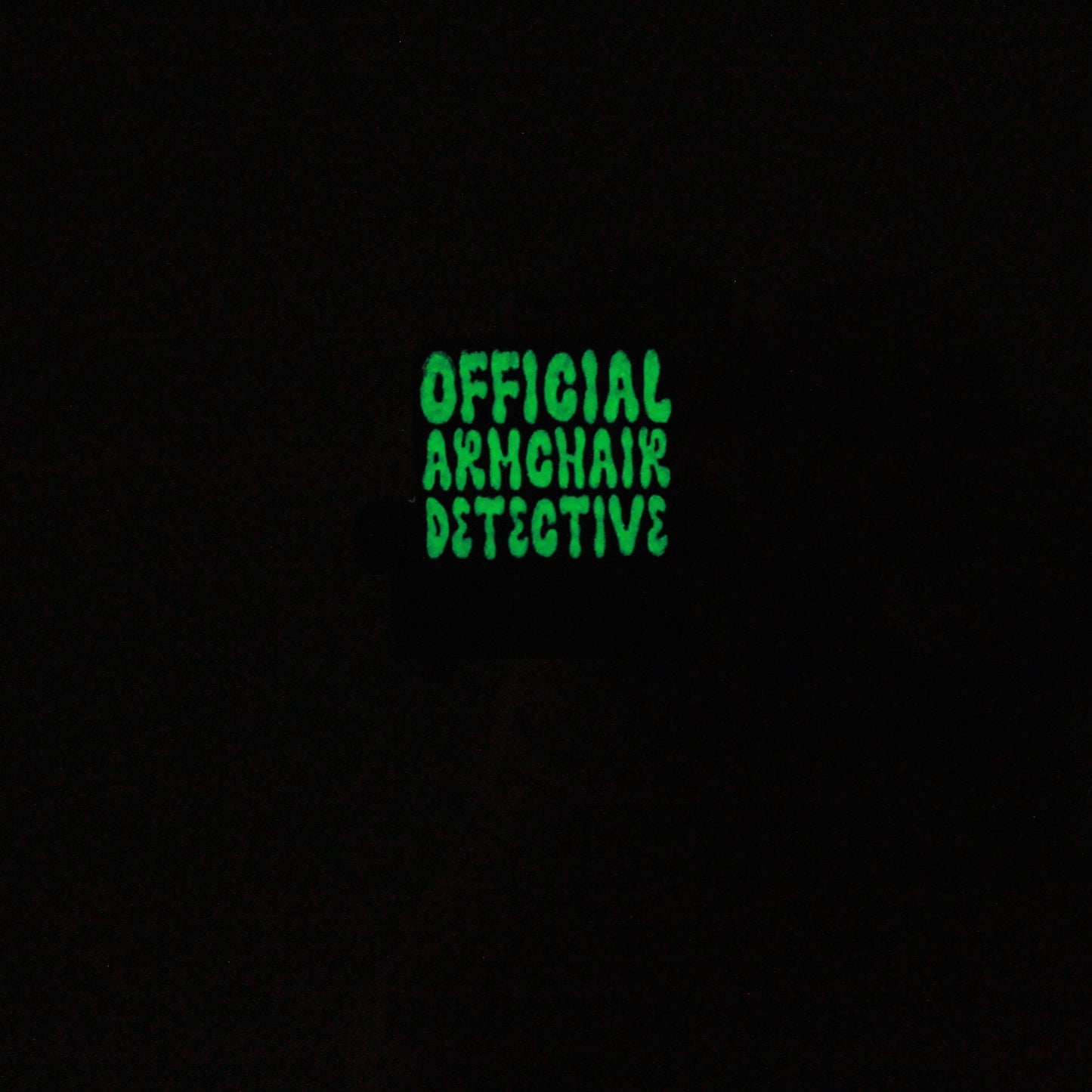 This photo shows the pin glowing in the dark. The lettering Official Armchair Detective glows a neon green in the dark after charging in light