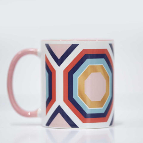 Hexagons in different colors inside of each other. Light pink on innermost hexagon, then white outline, mustard yellow, light blue, dark blue, orange ish red, then white. Mug has a light pink handle