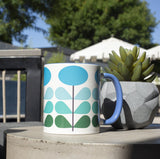 1950s mid century modern blue flowers on a white background coffee mug. Has ombre dark green to light blue leaves. Inside of mug is blue, and the handle is also dark blue. This photo showcases the mug next to a succulent