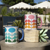 1950s mid century modern blue flowers on a white background coffee mug. Has ombre dark green to light blue leaves. Inside of mug is blue, and the handle is also dark blue. This photo also showcases this mug in pink. Photo is set outside next to succulents with the pink mug sitting higher than the blue mug