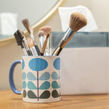 1950s mid century modern blue flowers on a white background coffee mug. Has ombre dark green to light blue leaves. Inside of mug is blue, and the handle is also dark blue. This photo shows the mug holding a variety of makeup brushes