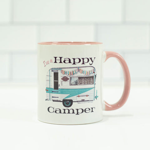 White mug with a white and teal 1950s trailer camper. Camper has a sign next to the door, aand fringe decor aling the top. Text says "I'm a happy camper"