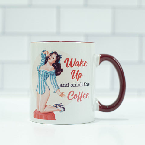 1950s style brunette pinup with a blue and white striped nightshirt on next to the words "wake up and smell the coffee" on a cream colored background. Mug has a maroon colored handle