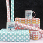White mug with a white and teal 1950s trailer camper. Camper has a sign next to the door, aand fringe decor aling the top. Text says "I'm a happy camper". This photo shows two of the mugs on top of wrapped presents in a 1950s mid mod print. One mug has wrapping paper set inside of it. On bottom right, shows another mug with pink mid mod flowers