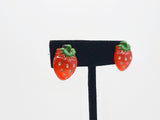 Red strawberry stud earrings with white seeds, and a green top. Earrings are shown on a black earring stand. This photo shows earrings from a side angle