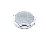 A round compact mirror with silver holographic glitter on one side of it. Compact has a small button to open the mirror