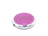 A round compact mirror with pink holographic glitter on one side of it. Compact has a small button to open the mirror