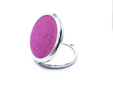 A round compact mirror with pink holographic glitter on one side of it. Compact has a small button to open the mirror. Photo shows mirror open and showcasing the glitter side of the mirror