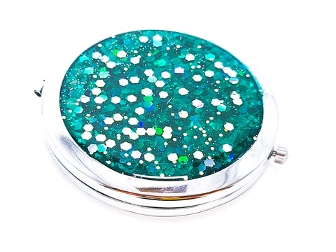 A round compact mirror with silver iridescent hexagon shaped glitter in a turquoise background on one side of it. Compact has a small button to open the mirror