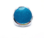 A round compact mirror with teal glitter on one side of it. Compact has a small button to open the mirror. Photo shows glitter side of mirror