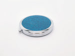 A round compact mirror with teal glitter on one side of it. Compact has a small button to open the mirror