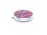 A round compact mirror with blue, yellow, pink, red, silver, and purple glitter on one side of it. Compact has a small button to open the mirror