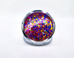 A round compact mirror with blue, yellow, pink, red, silver, and purple glitter on one side of it. Compact has a small button to open the mirror. Photo showcases the glitter side of the mirror