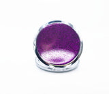 A round compact with Purple glitter on one side of it. Compact has a small button to open the mirror. Photo shows the purple glitter side of the compact