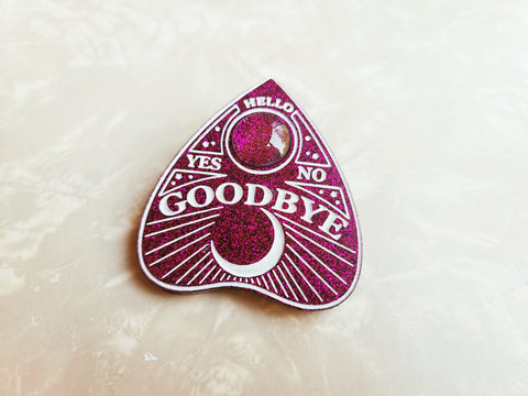 Glittery purple planchette hair clip with white lettering and detailing. Has a clear glass dome in the center of the planchette. Says: "hello" "Yes" "No" "Goodbye" with a quarter moon detailing along the bottom