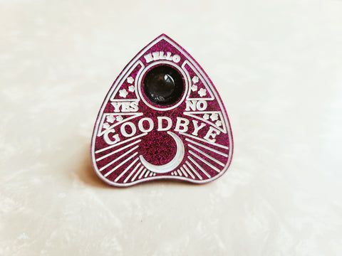 Glittery purple planchette phone grip with white lettering and detailing. Has a clear glass dome in the center of the planchette. Says: "hello" "Yes" "No" "Goodbye" with a quarter moon detailing along the bottom