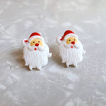 Photo shows the earrings up close. These earrings are of a White Santa with a red hat that has white fluffy edge and pom pom. Santa has a red nose, and a large white beard with a white mustache