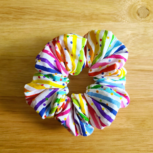 This photo shows the scrunchie against a wood background. Scrunchie is white with rainbow lines and rainbow confetti