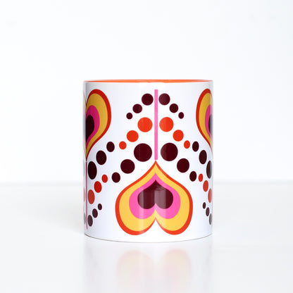 This mug has an orange, dark maroon, yellow, and pink coloring. Hearts have alternating colors inside of each other and are on a pink stem. Each heart flower alternates being upright and upside down with pink, maroon, and yellow dots as leaves flowing between each heart flower. This photo shows the mug's upside down heart. Mug handle, and inside coloring is orange